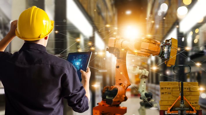 An engineer in a hard hat checking the automation of robot arms on a tablet in a modern industrial warehouse.