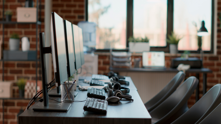 Fila de 3 puestos de trabajo vacíos con teclados y auriculares sobre el escritorio. Hay una ventana y una pared de ladrillo al fondo. 