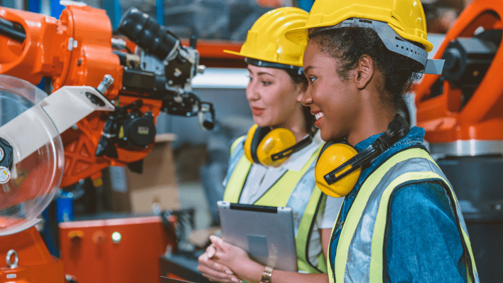 Mujeres con cascos interactuando con brazos robóticos en un entorno industrial.