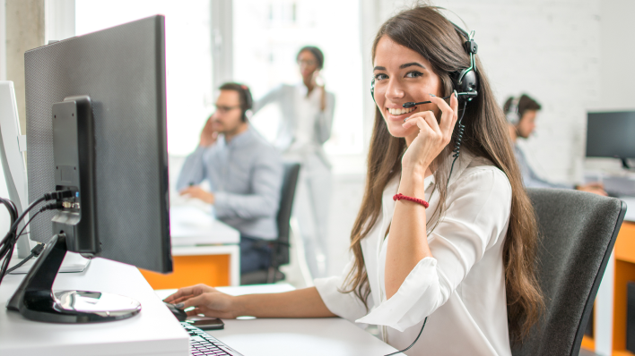 A modern call center setting focusing on a white woman with long dark hair. She wears a white blouse and a headset and smiles at the camera.