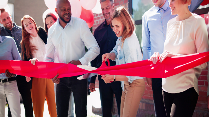 Ribbon-cutting ceremony: Mixed group of people in business-casual attire focused on red-haired white woman cutting a wide red ribbon with an oversize pair of scissors.