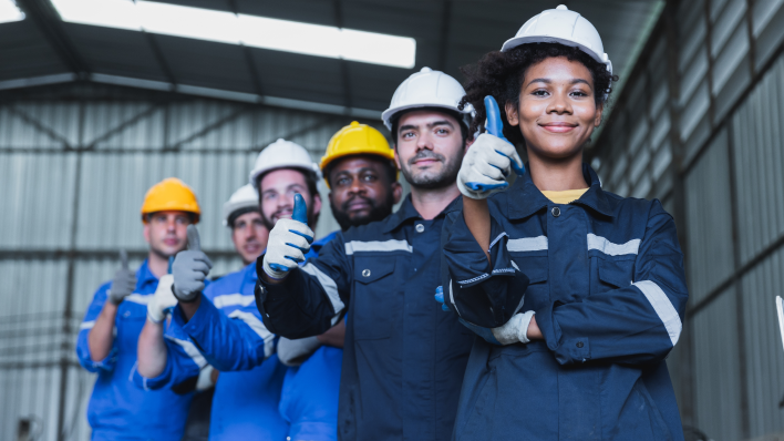 Una fila de 6 trabajadores multiculturales de una fábrica con cascos y guantes de trabajo. Todos miran a la cámara con el pulgar hacia arriba.