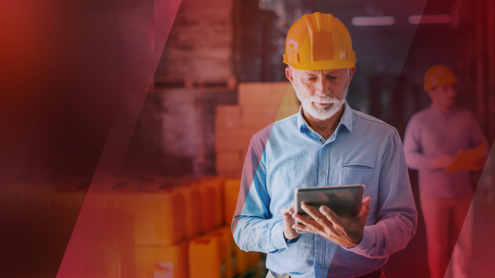 Man of approx. 60 years with gray beard wearing an orange hardhat and looking at a tablet in his hand.