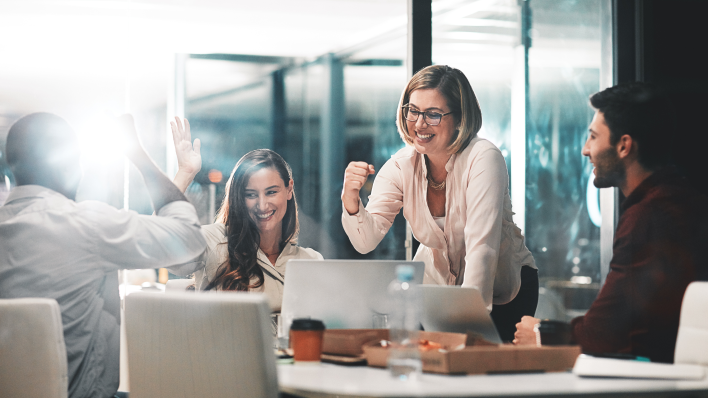 4 people (2 men, 2 women) in modern conference room. Smiling blonde woman is making a fist pumping motion and the woman to her right is about to high five man across the table.