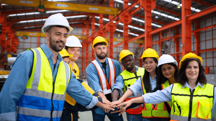 Seven multicultural male and female workers forming a semi-circle and reaching out to touch hands. All are wearing safety vests and hard hats.