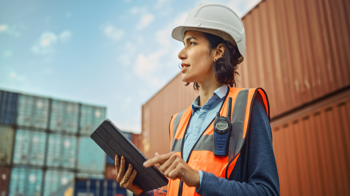 Mujer con casco en un entorno al aire libre. Tiene una tableta en la mano y mira hacia arriba.